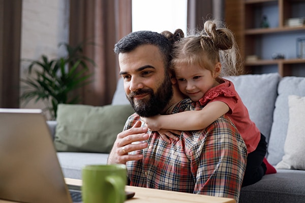 Man working on laptop with child hugging him and looking over his shoulder.